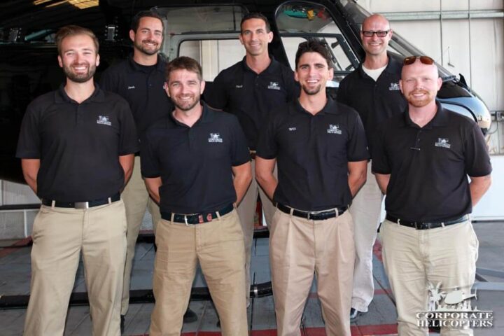 7 male filight instructors in uniform stand in front of a helicoper