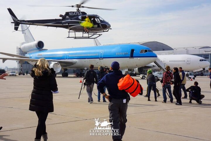A helicopter hovers over an airplane. A woman in a yellow dress sites on the open helicopter door.