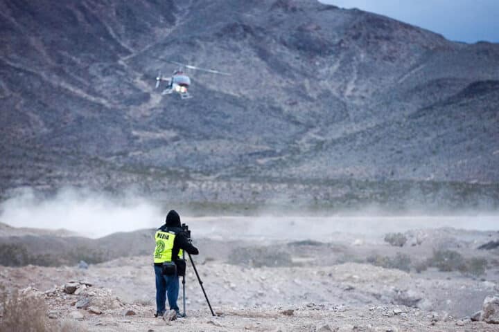 Man in desert filming race car and helicopter above