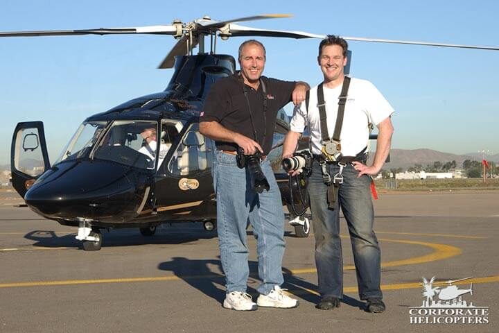 2 Aerial photographers with cameras stand in front of helicopter