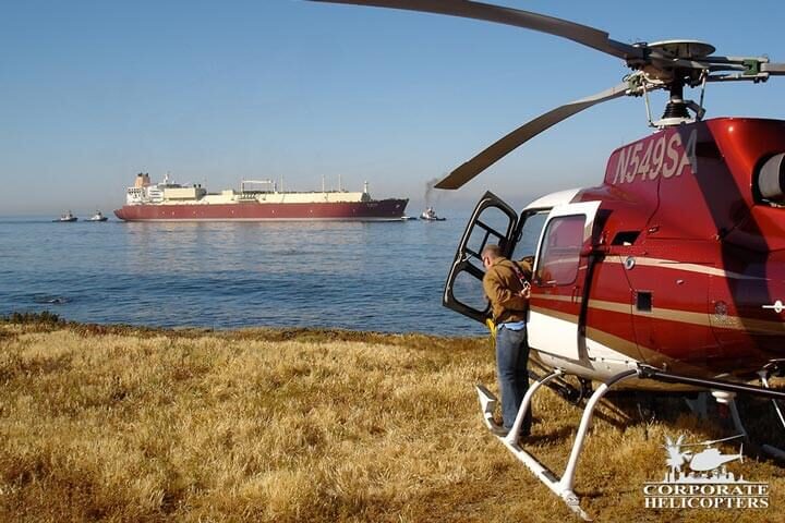 A helicopter landed next to the ocean, a barge and boats are in the ocean