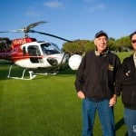 Two men stand in front of a helicopter on a golf course