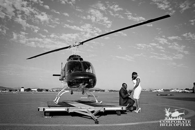 A man on one knee with his bride, next to a helicopter